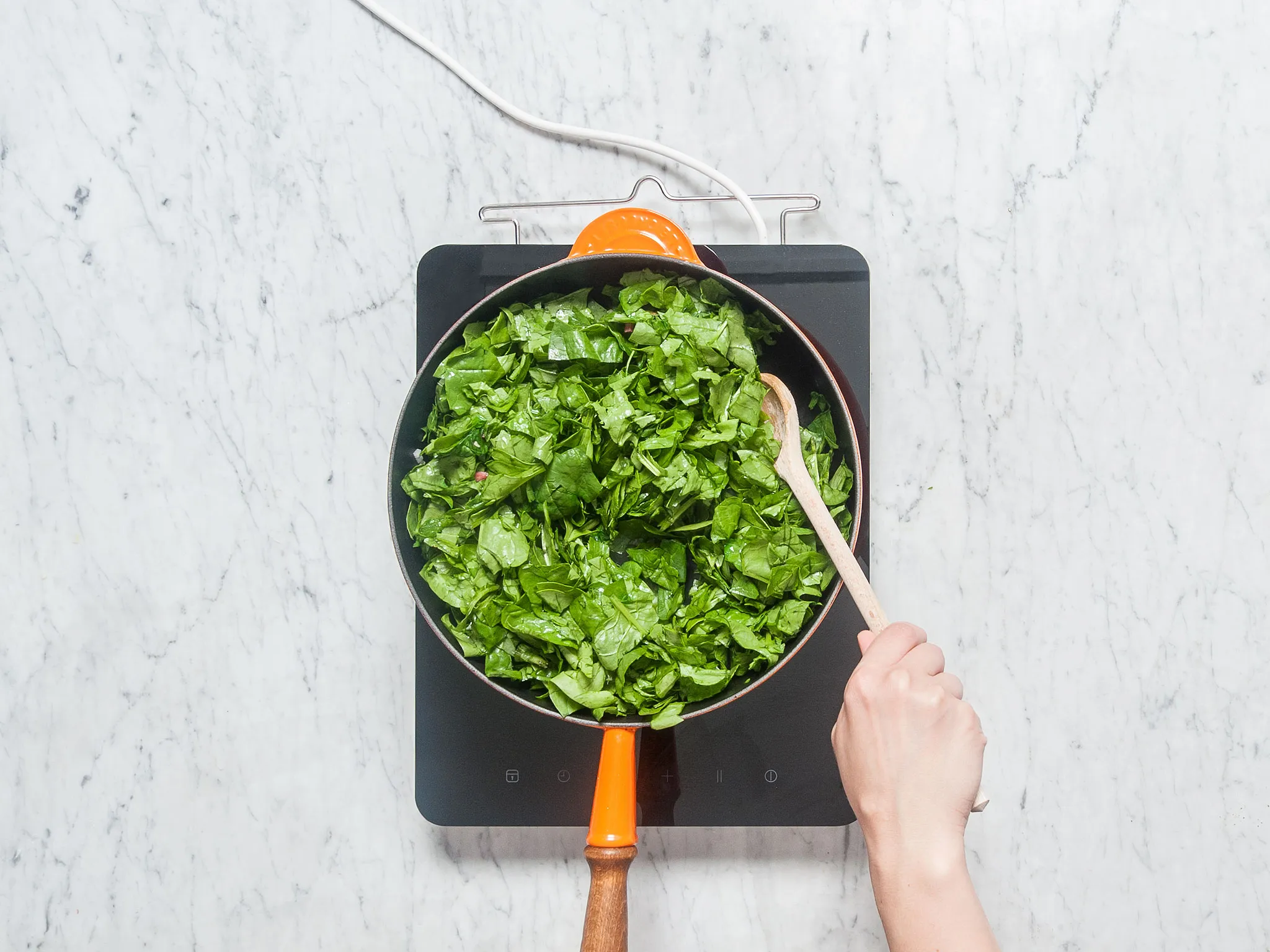 Person stirring spinach in a pan with a wooden spoon on an induction cooktop.