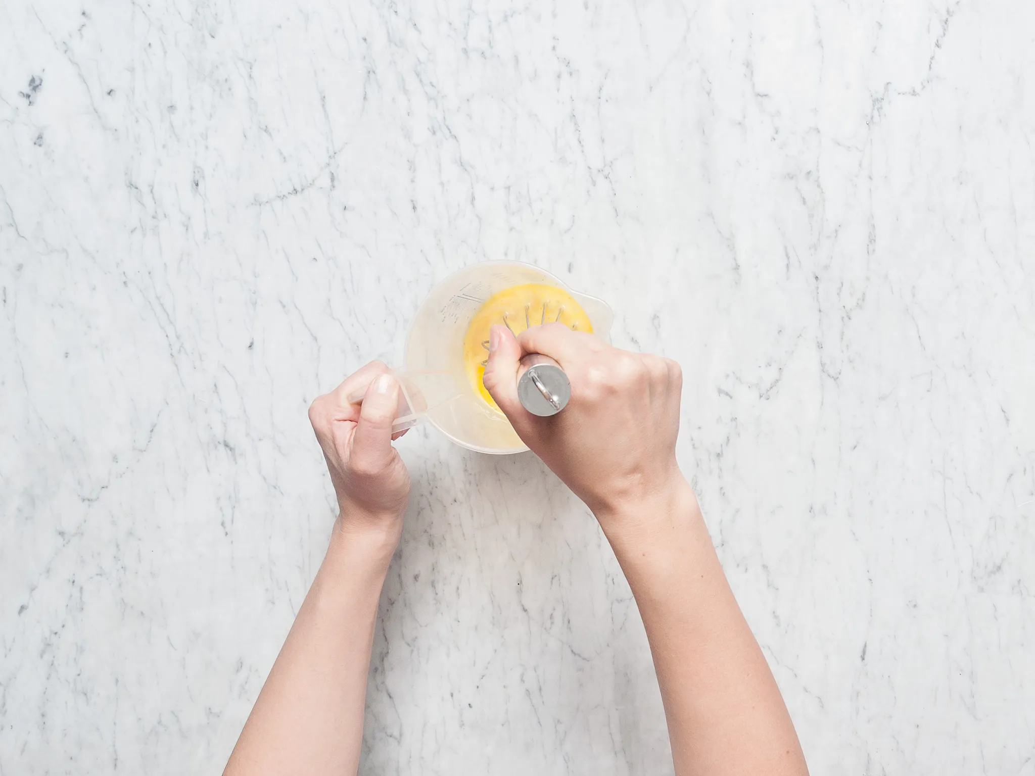 Hands whisking yellow batter in a measuring cup on a marble counter.