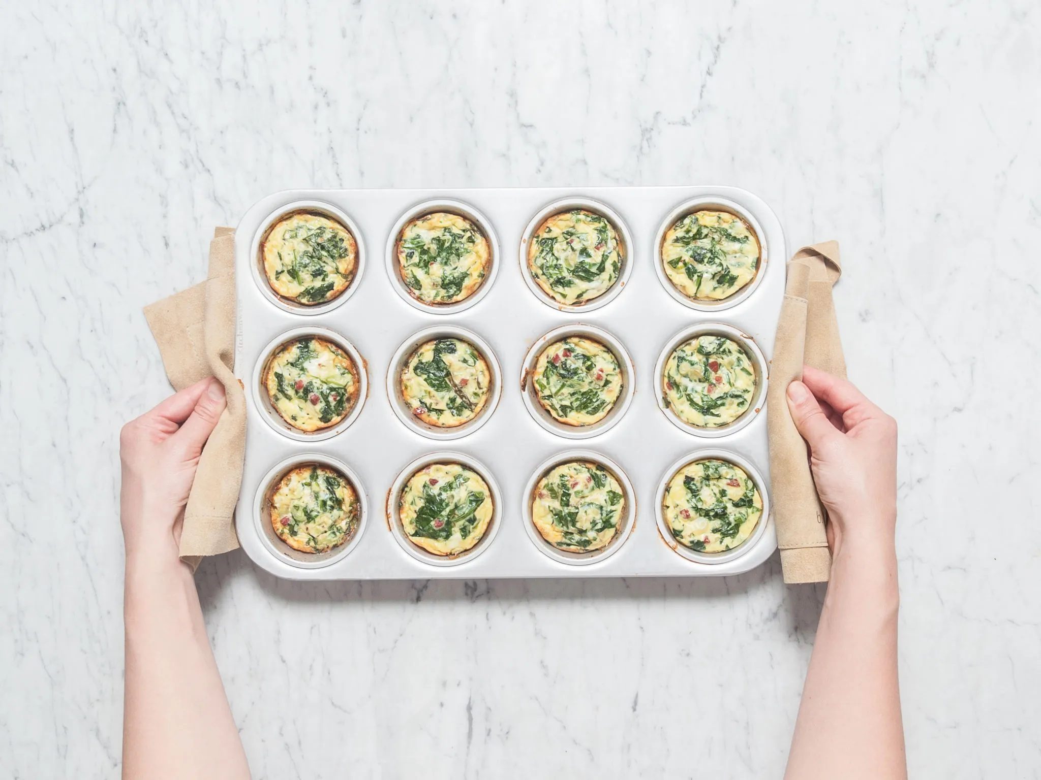 Hands holding a tray of mini spinach quiches on a marble countertop.