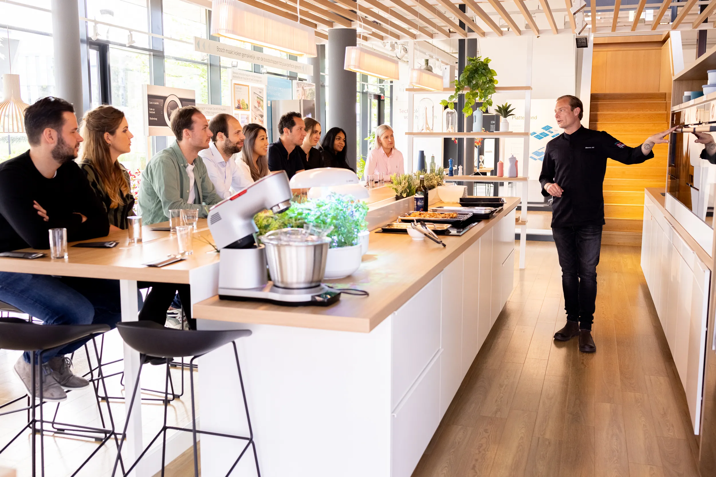 alt="Groep van mensen verzameld in een moderne, open kantoorruimte met een keuken en planten."