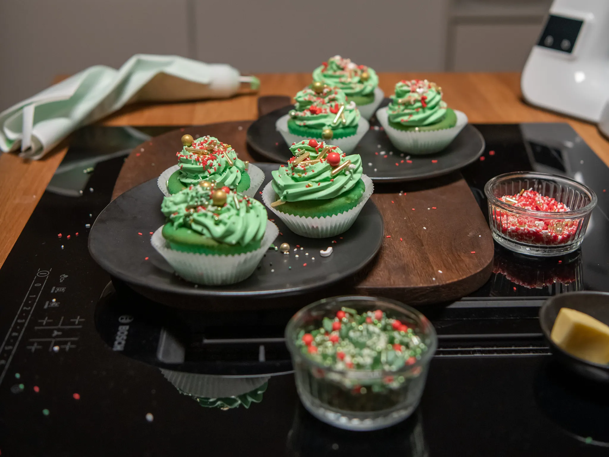 Decorative green cupcakes on black plates with sprinkles on a wooden board in a kitchen.