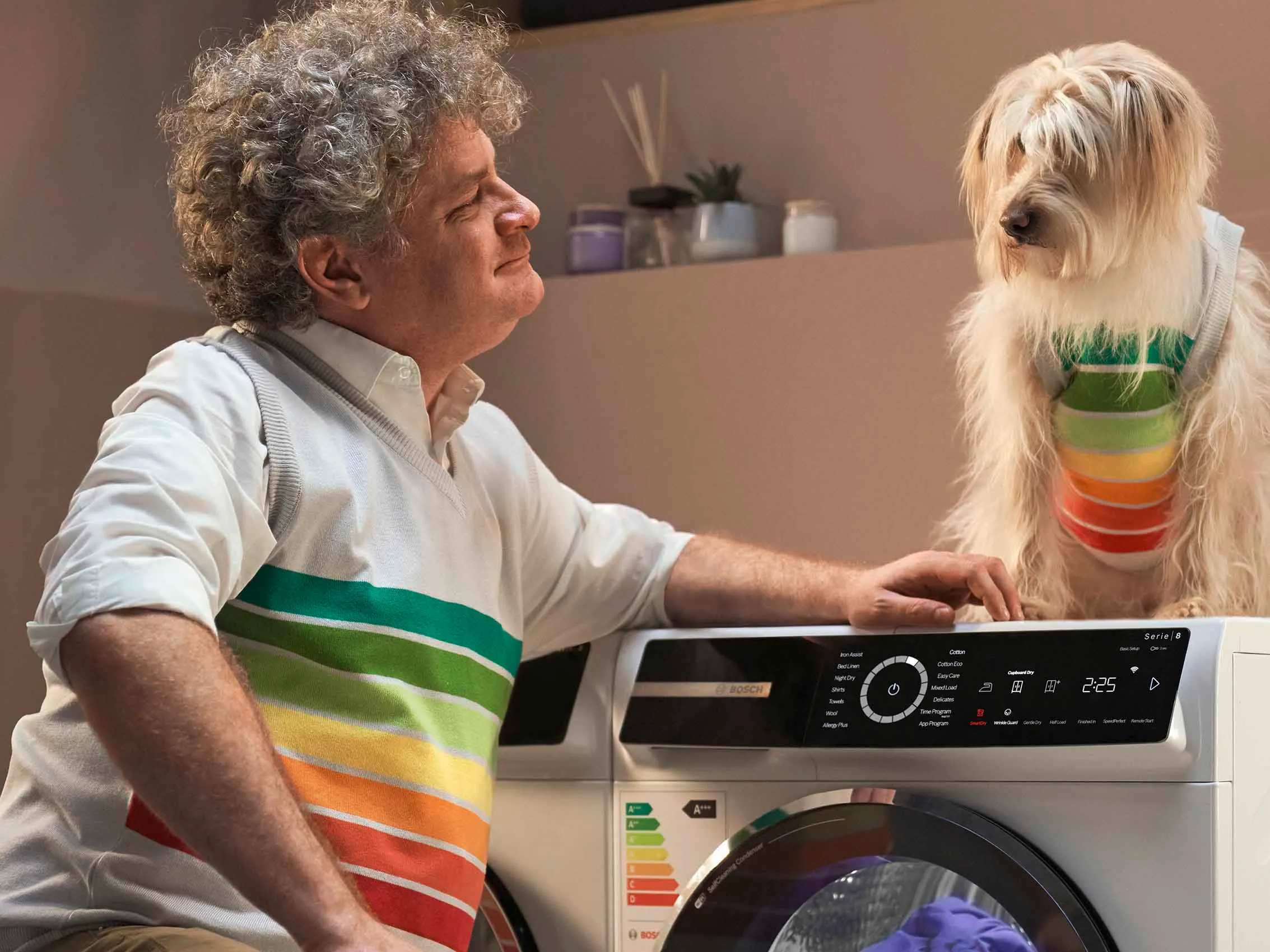 Mies nojaa kuivausrumpua vasten, ja katsoo hymyillen koneen päällä istuvaa koiraa.