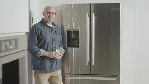 Older man with gray beard standing next to Bosch stainless steel refrigerator.