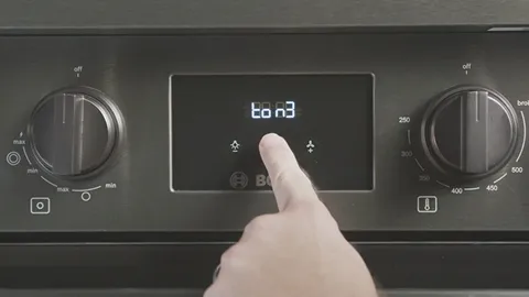 Oven control panel with digital display, knobs, and a hand pressing a button.