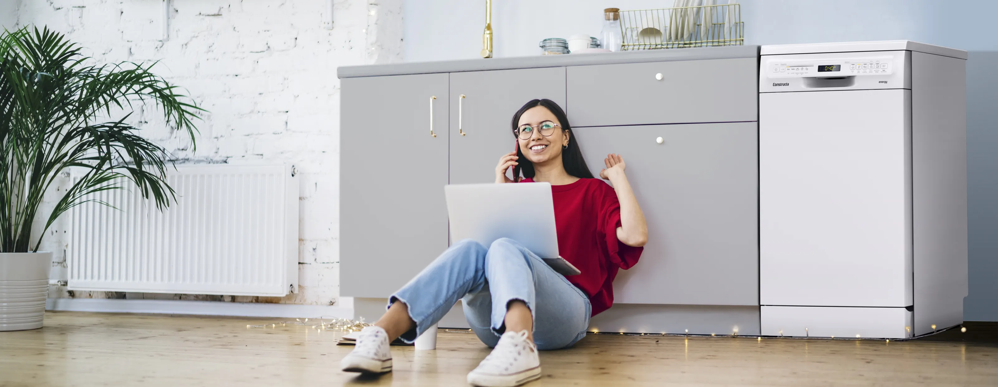 Eine junge Frau telefoniert und sitzt mit ihrem Laptop auf dem Boden neben einem Constructa Geschirrspüler.