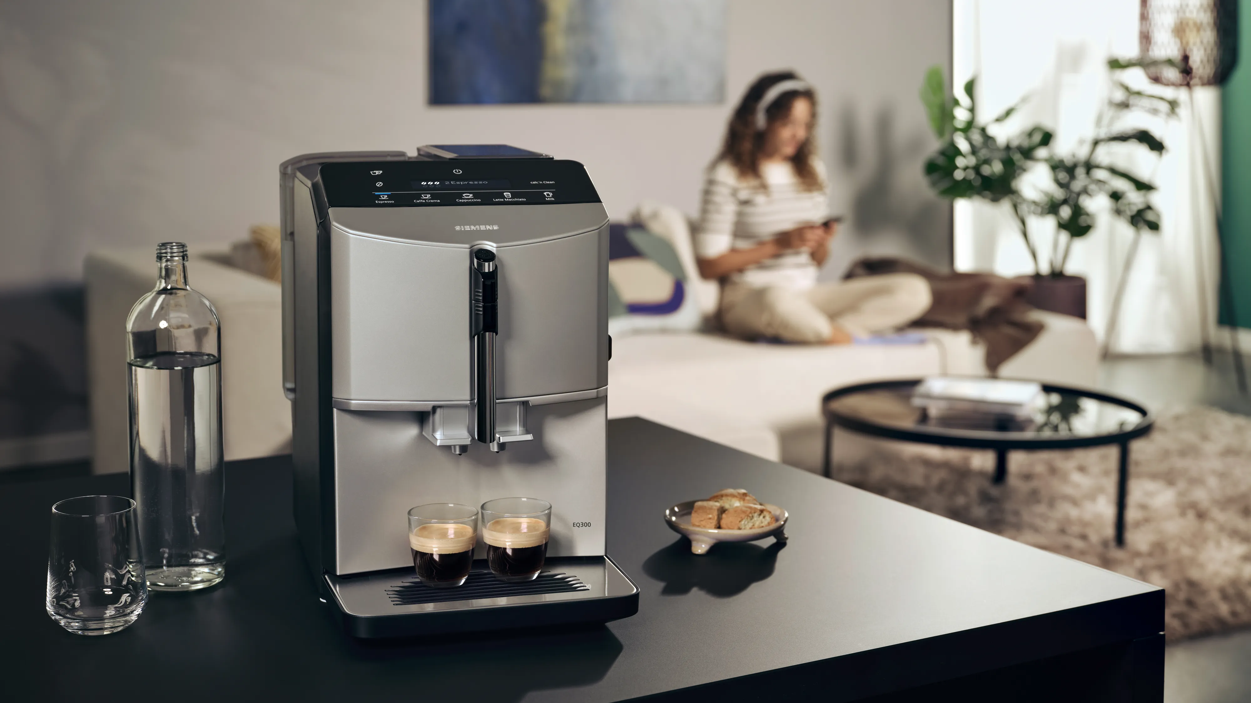 EQ300 fully automatic espresso machine on a kitchen island is preparing two espressos, in the background a woman on a sofa.
