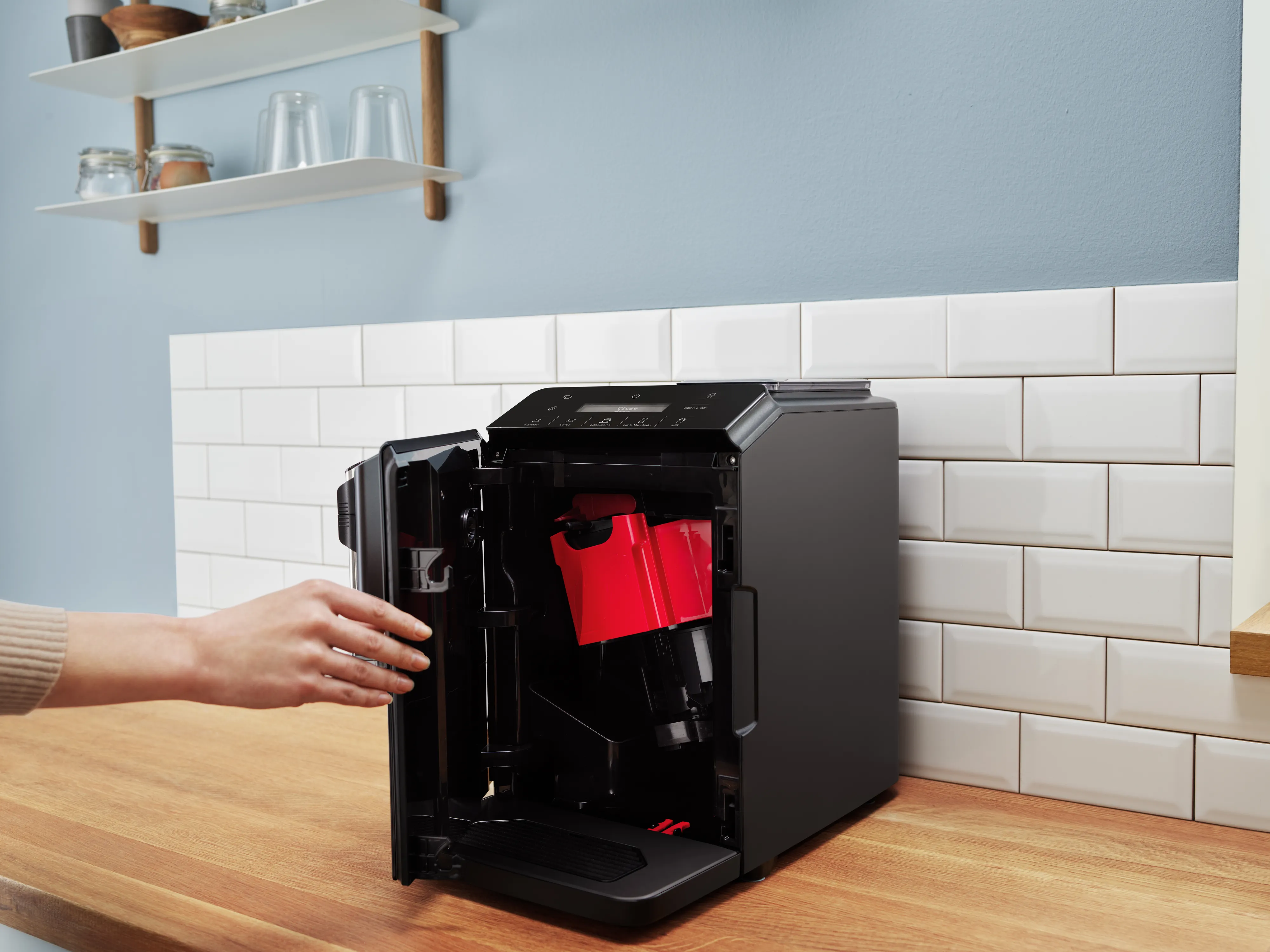 A person opens a Bosch coffee machine on a wooden counter with tiled backsplash.