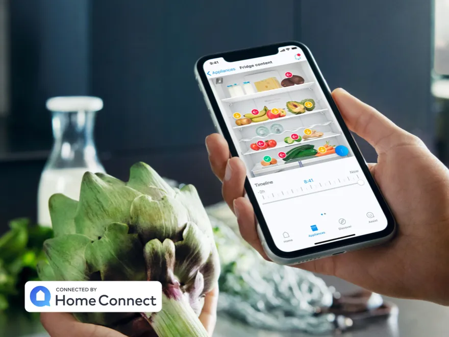 Una persona tiene in mano un carciofo e uno smartphone che mostra l'app con la visualizzazione dell'interno del frigorifero, connessa tramite Home Connect.