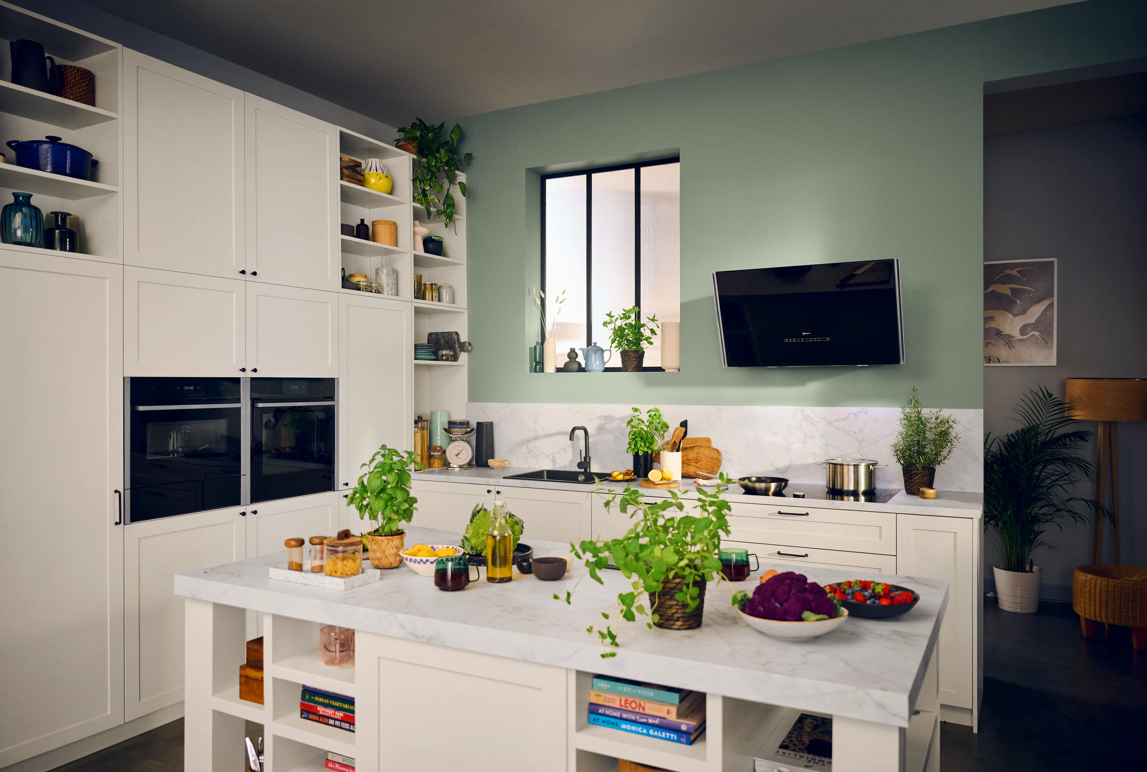 Cuisine claire et moderne avec des armoires blanches, un îlot en marbre, des plantes et des ingrédients colorés disposés pour la cuisson, avec une fenêtre et une télévision.