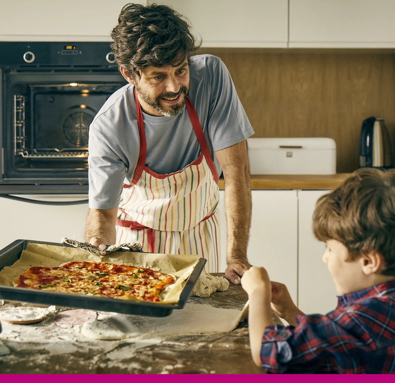 Eine Person in einer gestreiften Schürze präsentiert ein frisch gebackenes Pizza einem Kind, umgeben von Mehl und Küchengeräten in einer modernen Küche.