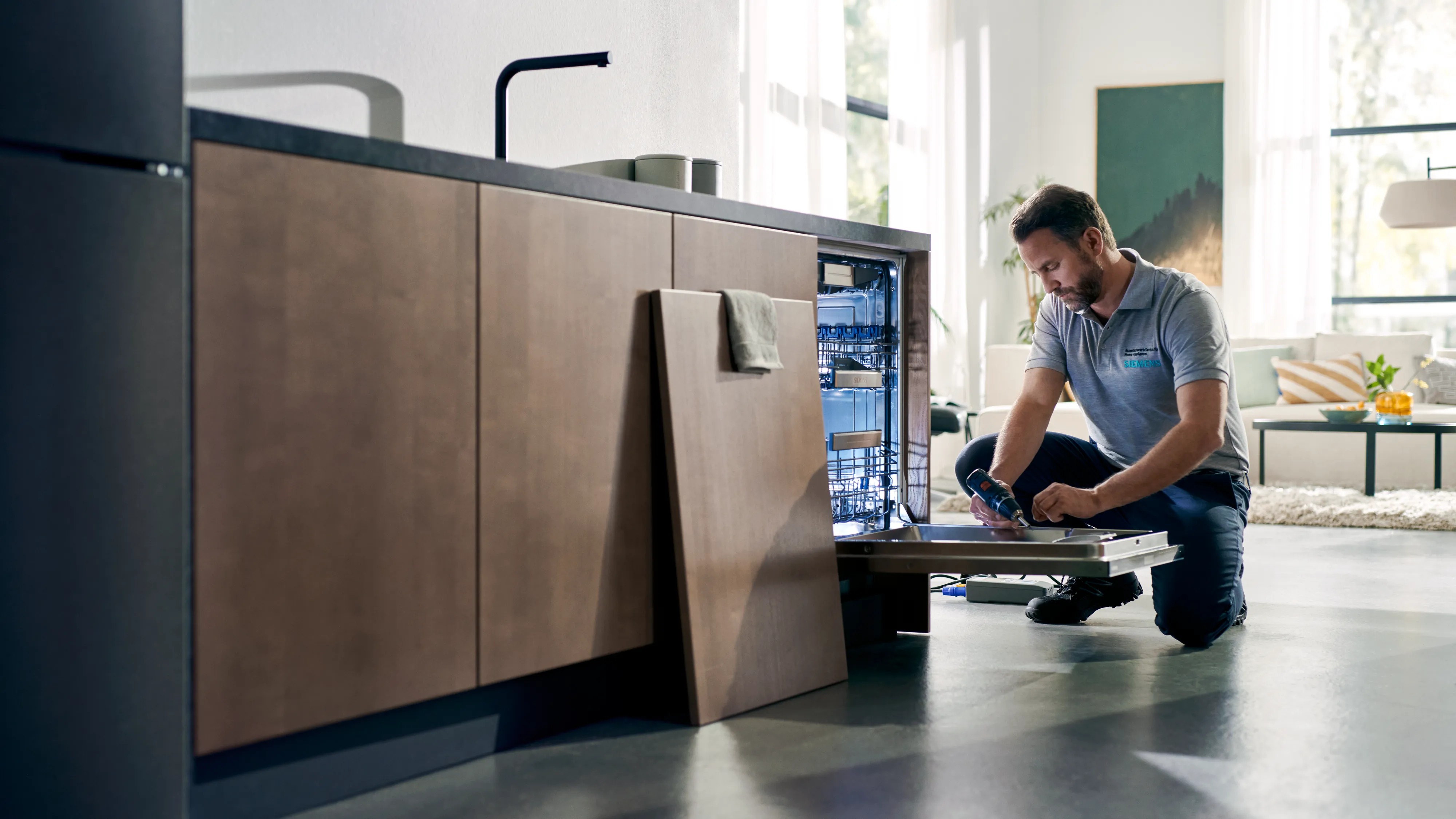 alt-A man in a grey uniform is kneeling next to a dishwasher with a screwdriver in his hand in a modern wooden kitchen.