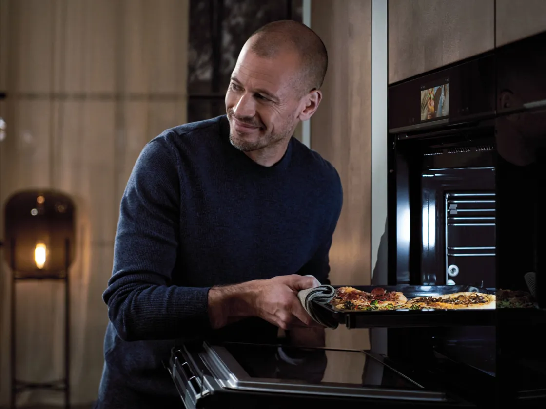 Man in dark sweater removing pizza from oven in kitchen.