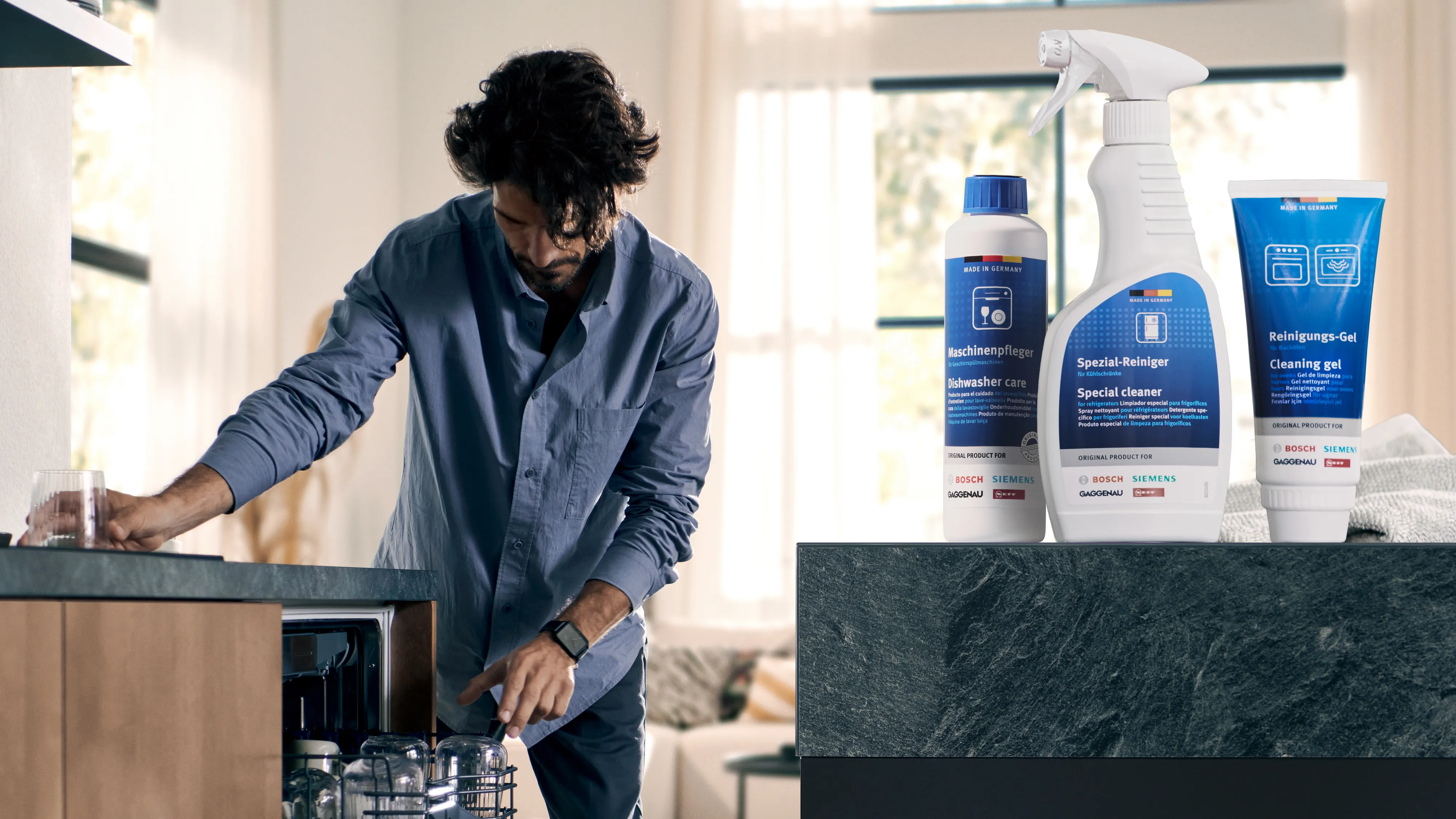 alt-A man in a blue shirt unloading a dishwasher and various cleaning products placed in the front corner of the picture.