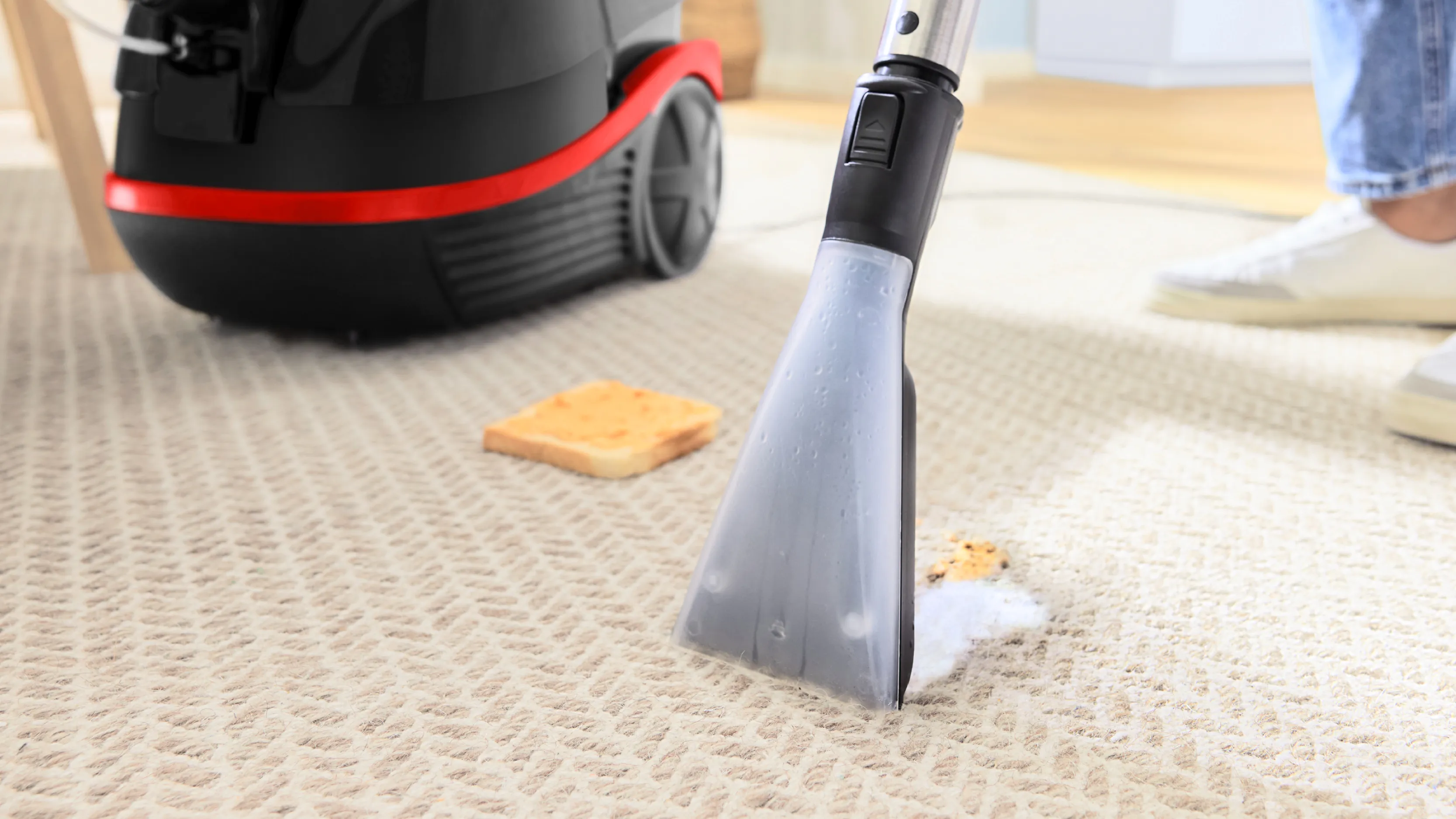 Vysavač čistí drobky na koberci s kouskem chleba viditelným na podlaze.