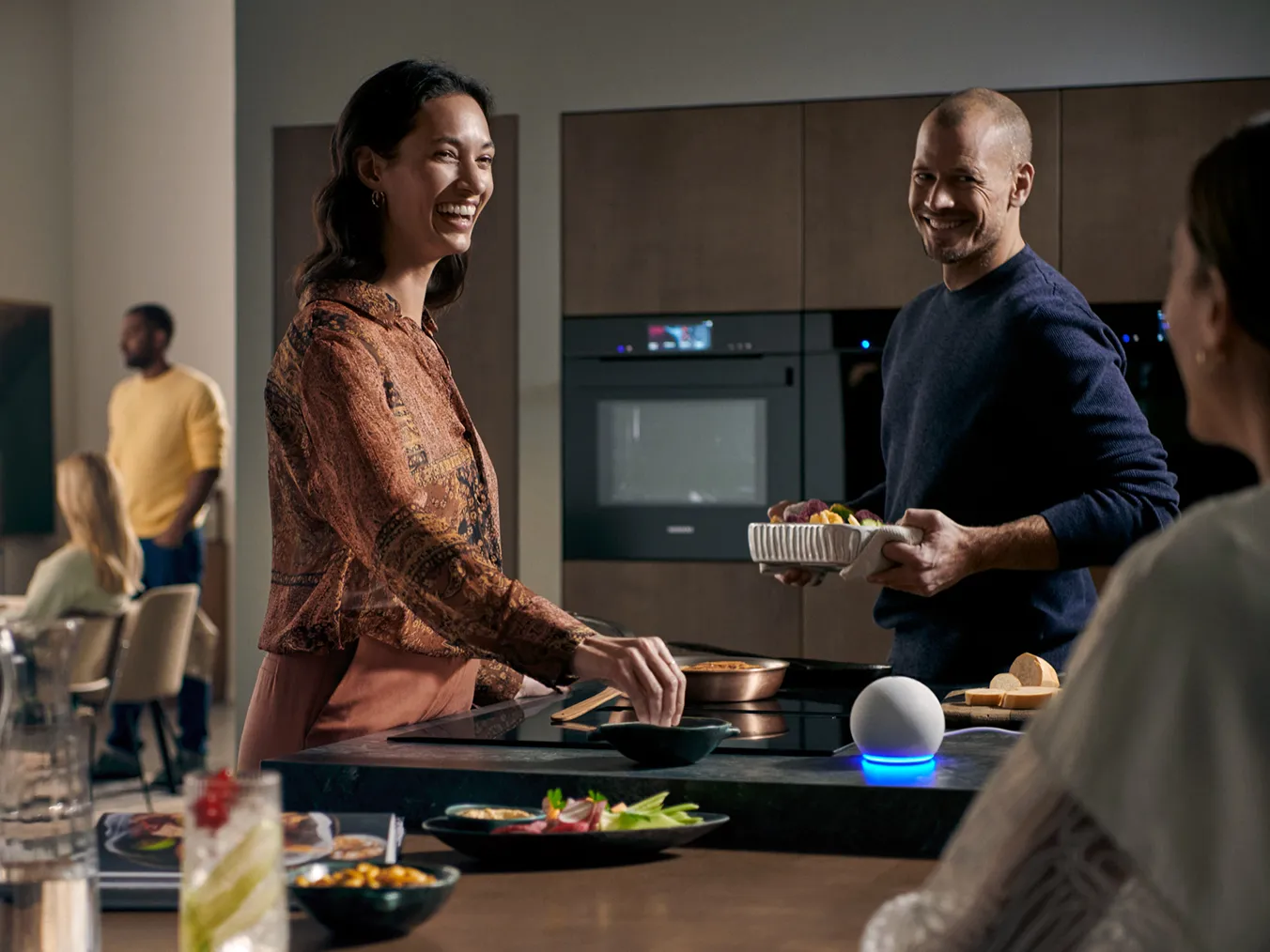 A smiling man and woman are happily preparing a meal together in a modern, stylish kitchen, surrounded by friends. An Amazon Echo Dot sits on the kitchen island, suggesting a smart home environment.