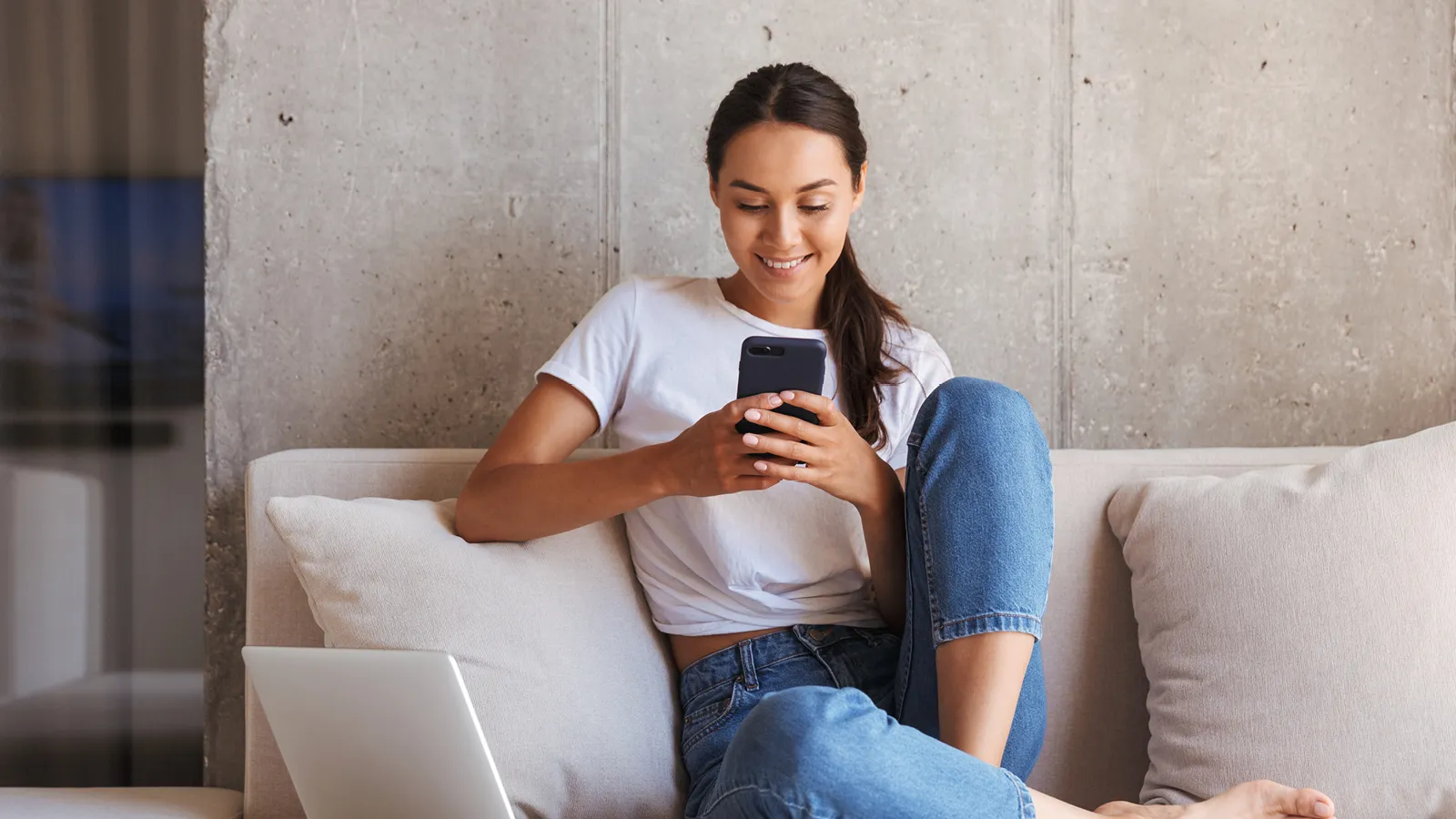 Young woman sitting on couch, using smartphone, laptop on lap, casual attire.
