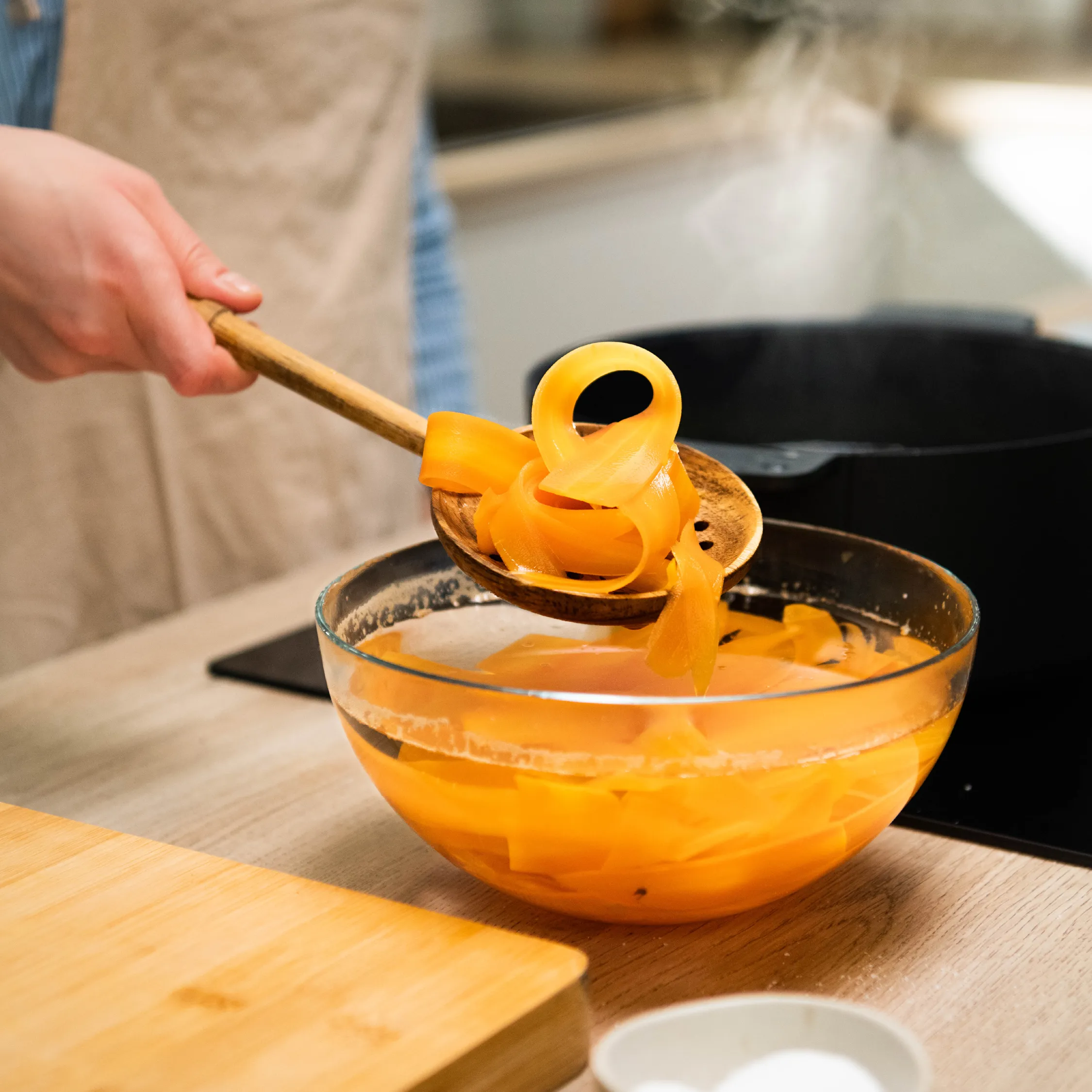 Close-up of a person’s hand holding a wooden slotted spoon lifting curly orange vegetable ribbons from a glass bowl of water on a kitchen counter, with