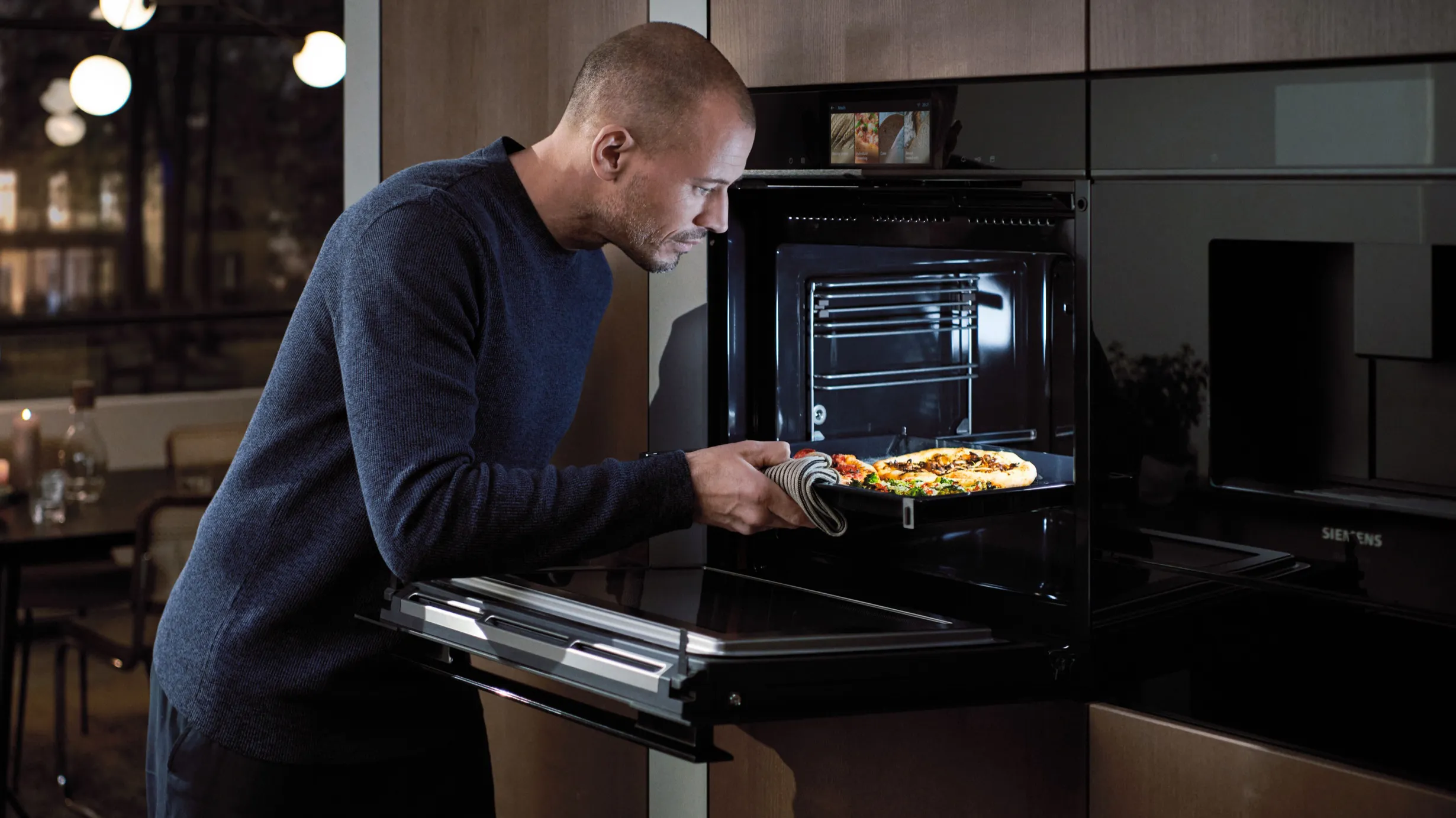 Man in dark sweater removing pizza from oven in modern kitchen at night.