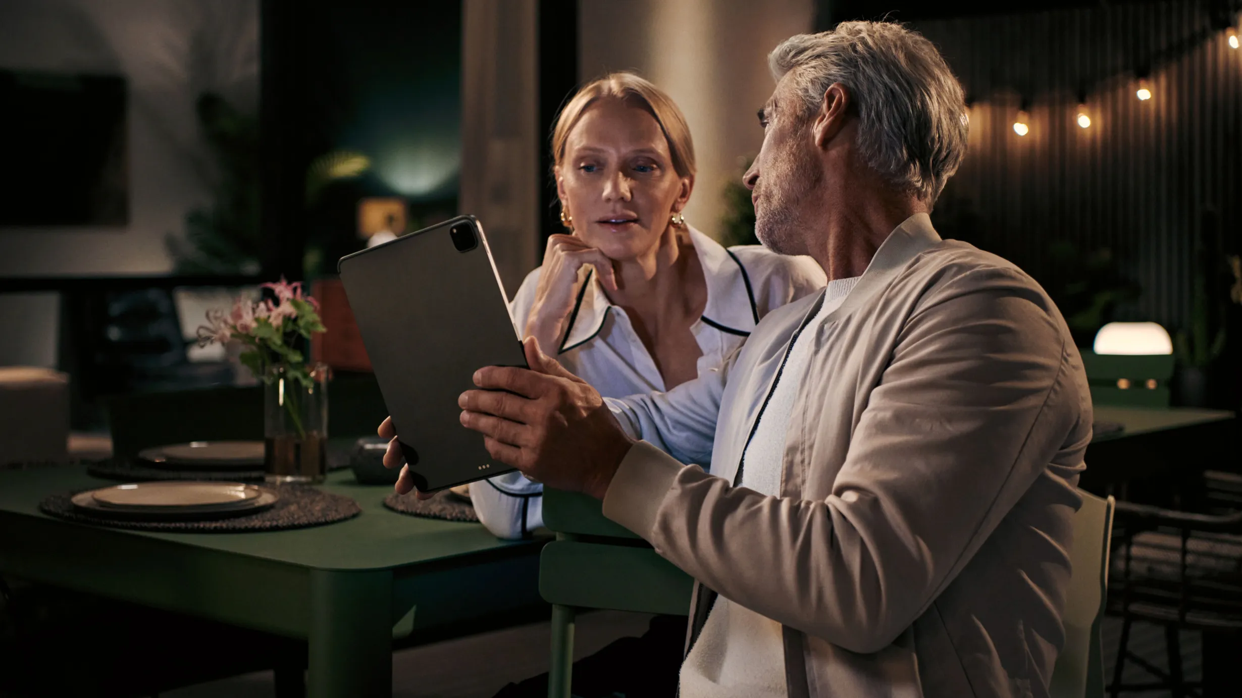 Older couple sitting at table, looking at tablet, intimate conversation in dimly lit room.