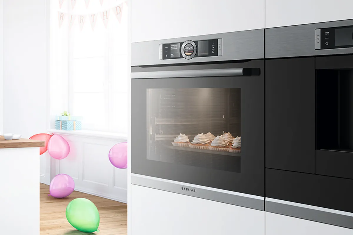 A modern kitchen with a Bosch oven baking cupcakes, and a birthday celebration setup with balloons and a gift box in the background.