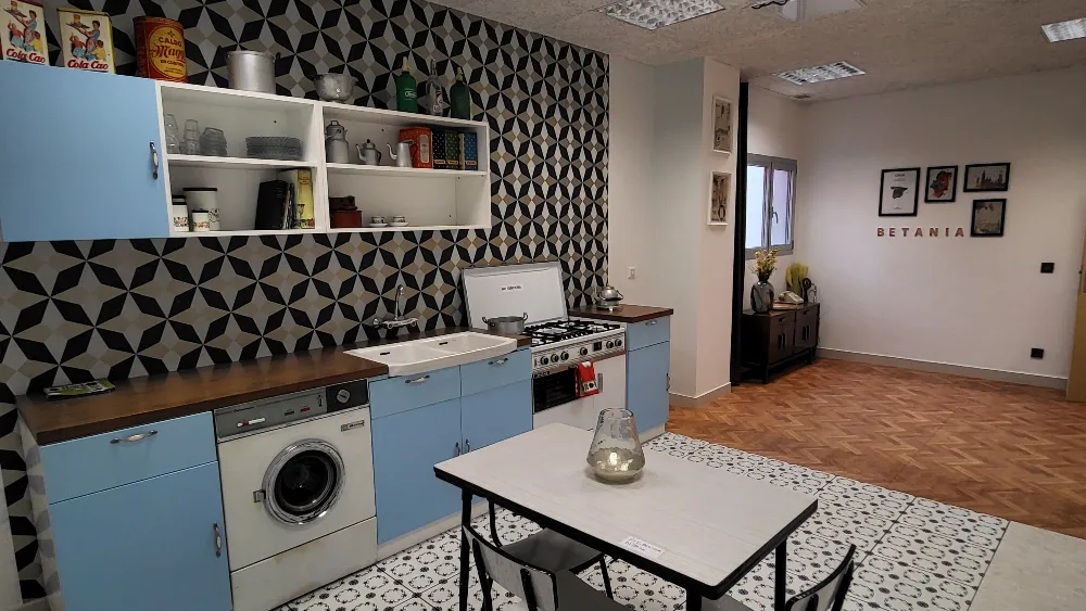 Cocina de estilo retro con azulejos estampados, electrodomésticos azules, mesa de comedor y estanterías que muestran la decoración. 