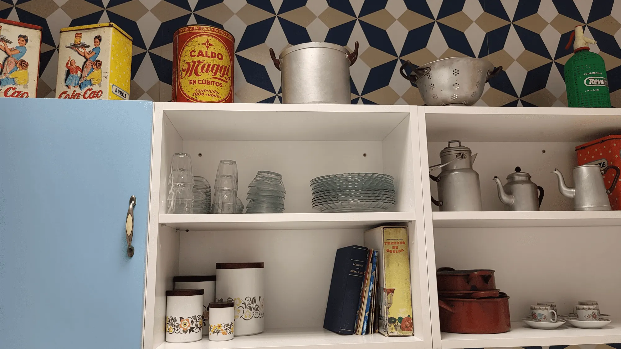 Estantes de cocina vintage con latas, platos y utensilios de cocina retro contra una pared estampada. 