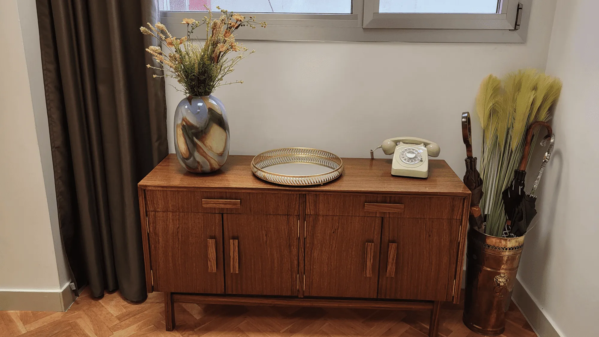 Credenza de madera con jarrón de flores secas, teléfono rotativo antiguo, bandeja decorativa y paraguas. 
