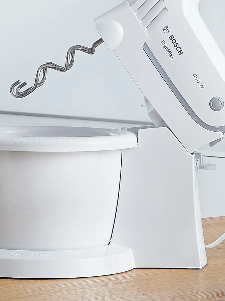A Bosch mixer with whisks and a food processor, beside a bundt cake and strawberries, on a wooden countertop in a bright kitchen.