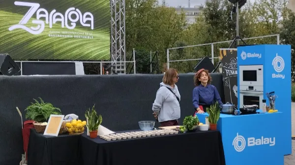 Demostración de cocina con mesa negra, ingredientes frescos, productos Balay y fondo de escenario "Zaragoza". 