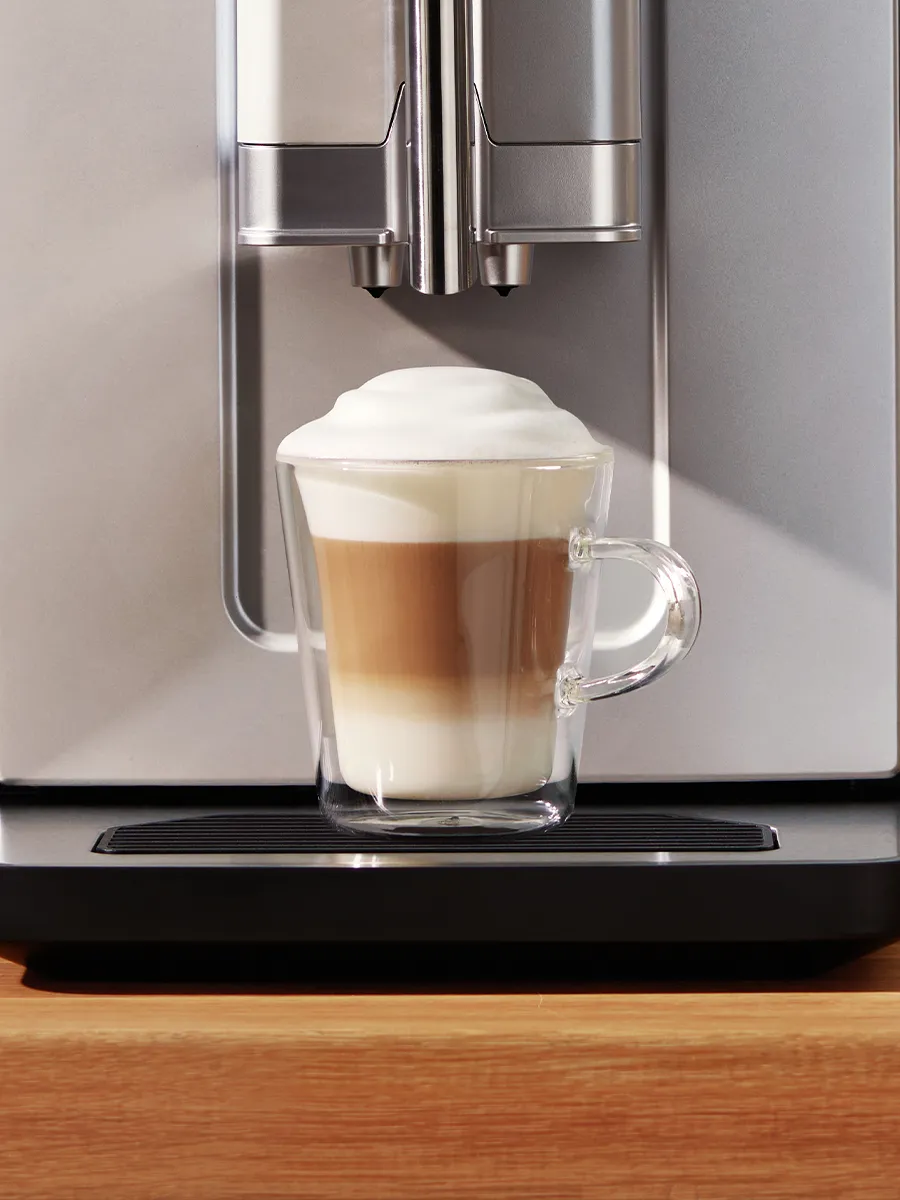 A glass cup with layered coffee, milk, and froth sits on a coffee machine's drip tray, showcased against a sleek silver appliance.