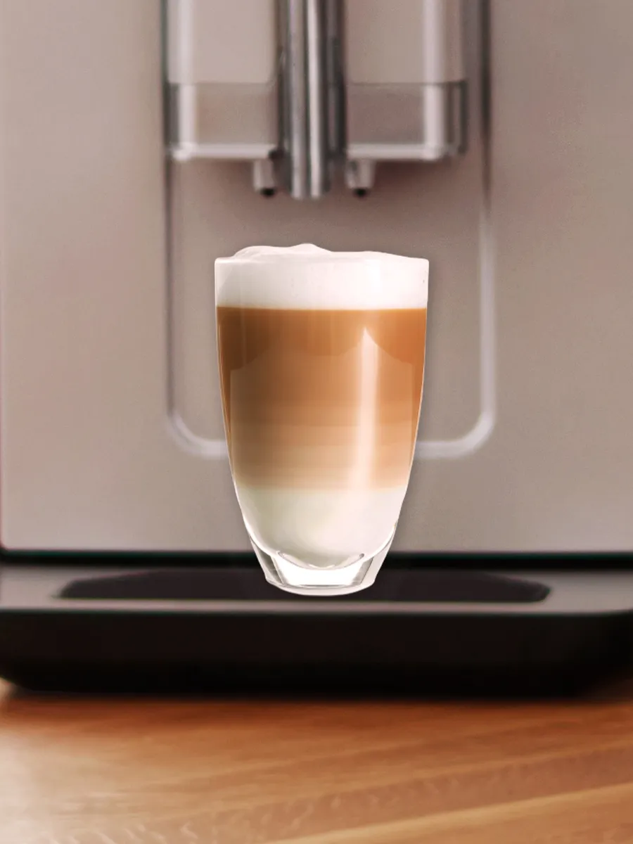 A layered latte in a clear glass, featuring coffee, milk, and froth, positioned in front of a sleek coffee machine.