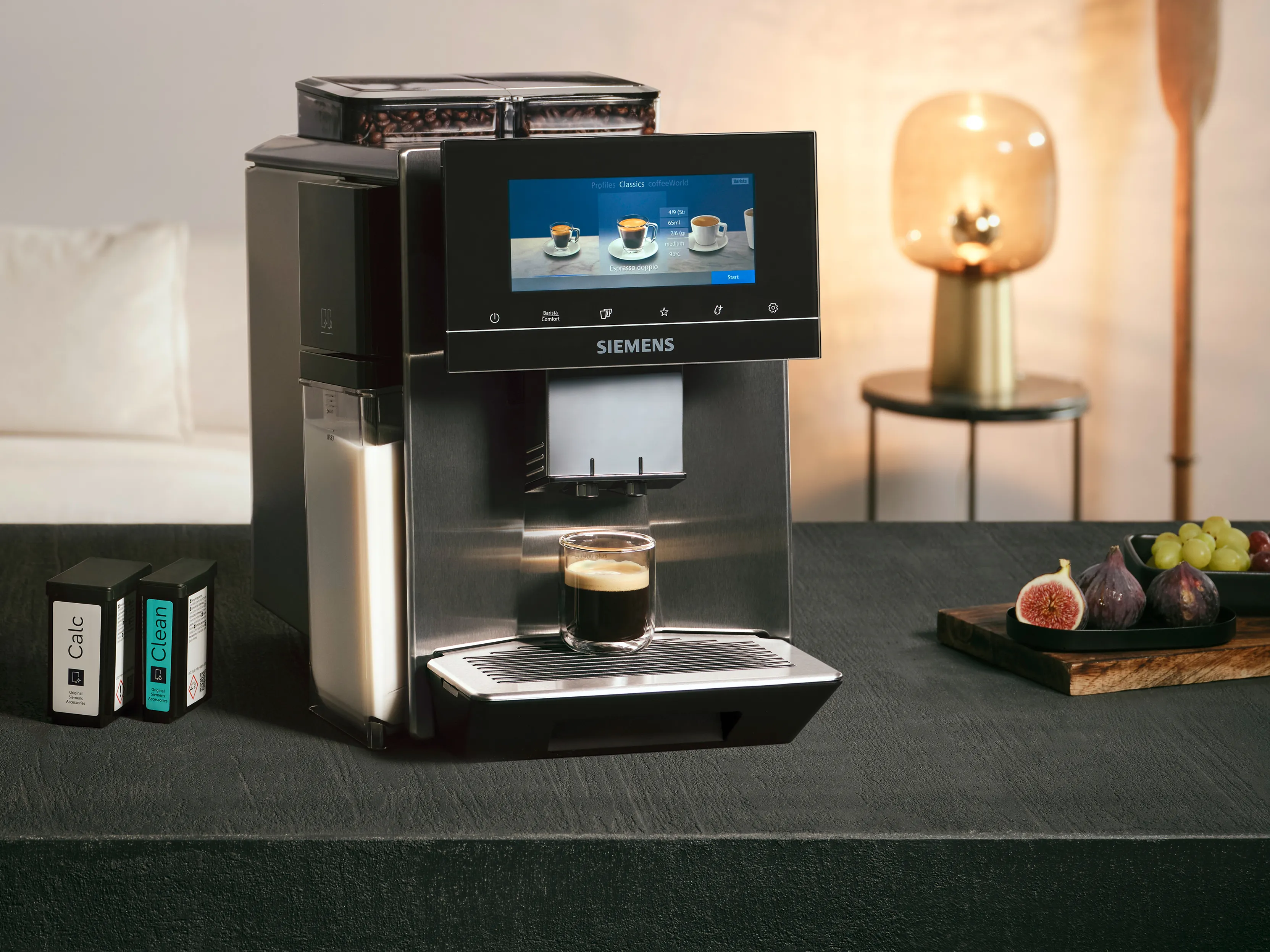 EQ900 plus fully automatic espresso machine stands on a black table, next to it a wooden plate with fruit, a kitchen towel and, on the left, cleaning and descaling cartridges