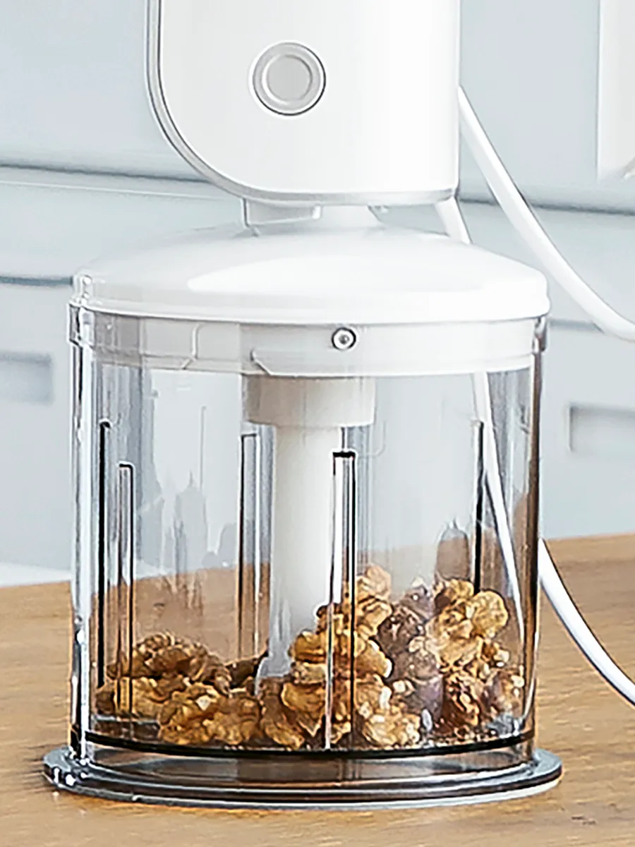 A black handheld mixer beside a transparent bowl, featuring mashed potatoes, walnuts, and a wooden spoon in a modern kitchen setting.
