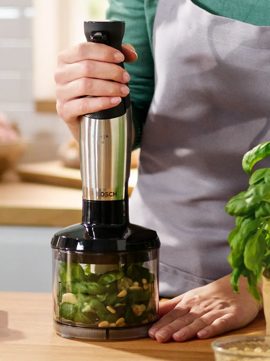 Hand using a Bosch hand blender to mix nuts in a clear container, with coconut-covered treats and kitchen items in a bright, modern setting.