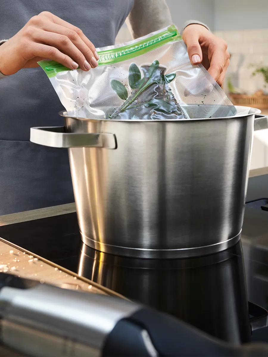 Hands placing a resealable plastic bag with fresh herbs into a large stainless-steel pot on an induction cooktop; person wearing a gray apron, with a blurred immersion blender in the foreground.