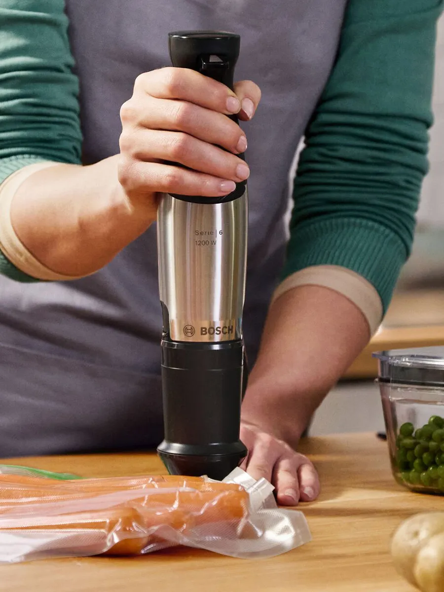 Close-up of hands using a stainless-steel Bosch Series 6 immersion blender (1200 W) held vertically over a vacuum-sealed bag of carrots on a wooden countertop; a container of green peas and some potatoes sit nearby, and the person wears a green-sleeved shirt and gray apron.