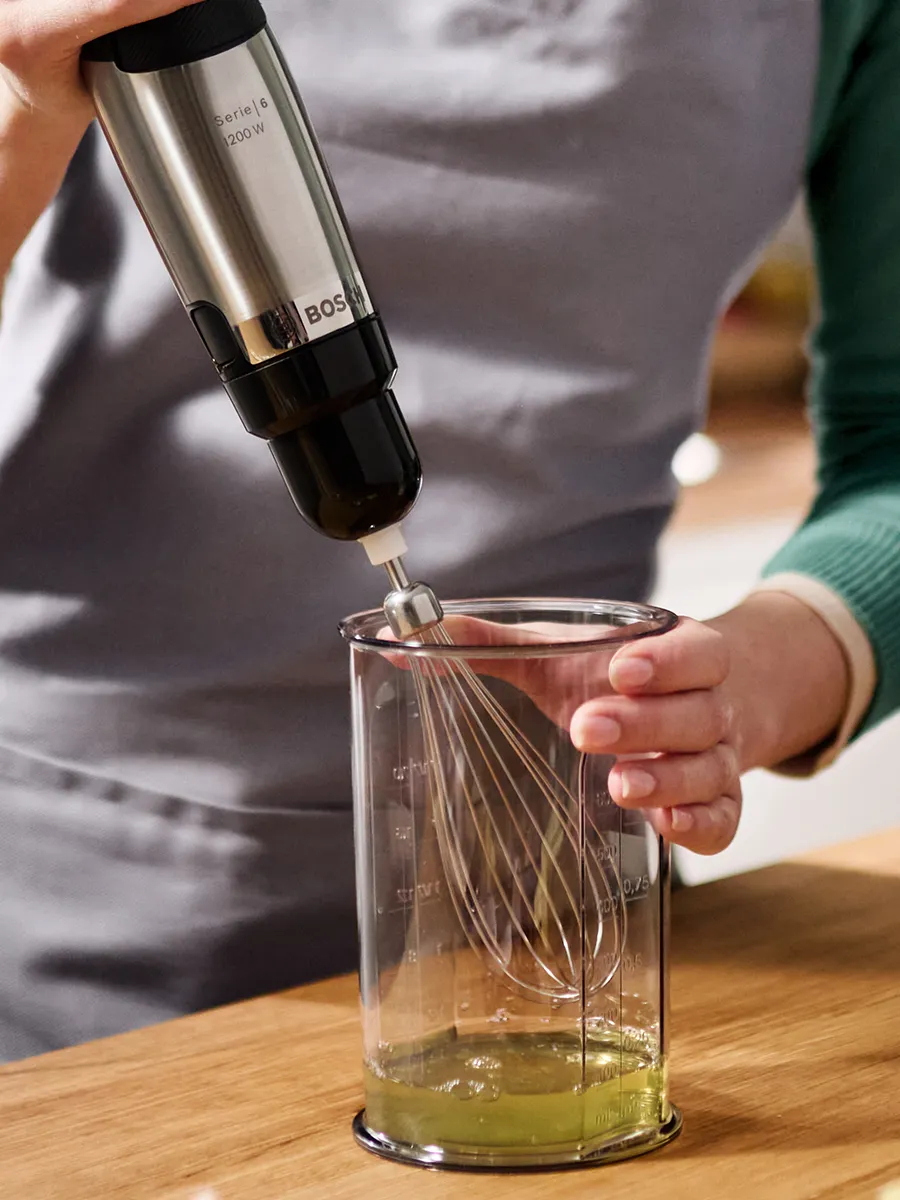 Someone is about to use a hand blender with a whisk attachment to beat egg whites in a beaker.