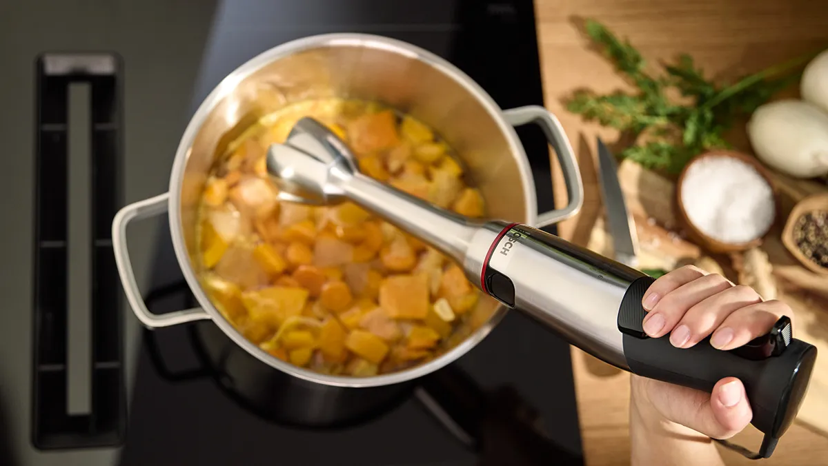 A hand holds an immersion blender over a pot of freshly cooked vegetables, ready to make smooth soup.