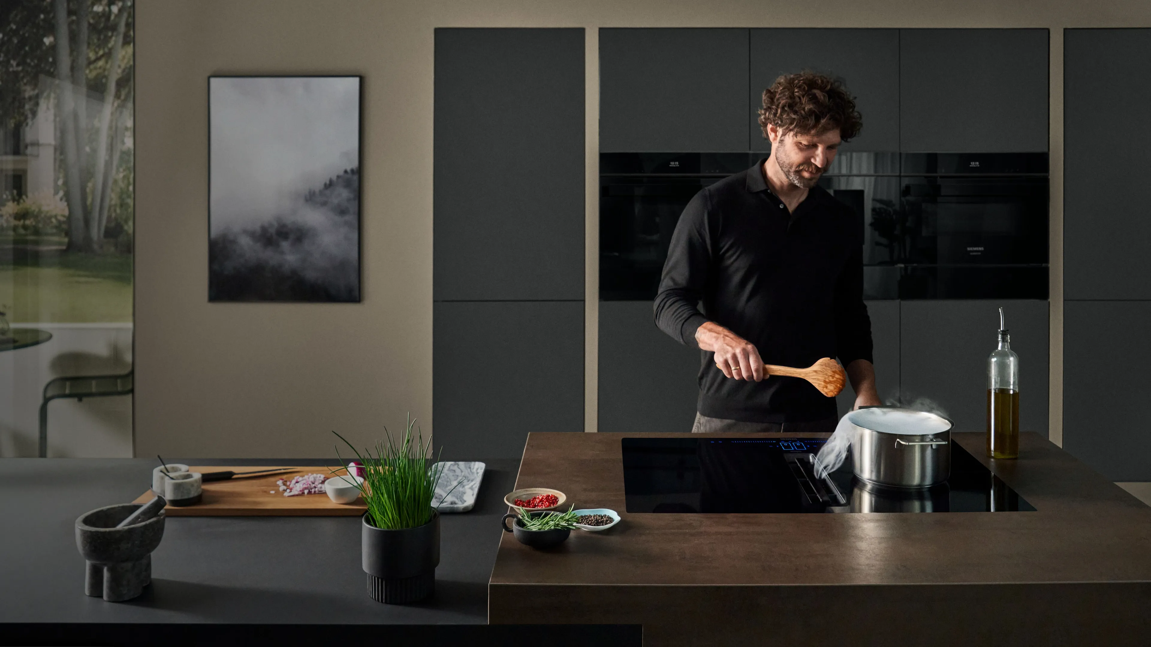 Kocken förbereder måltid i modernt kök med mörk inredning och stora fönster.