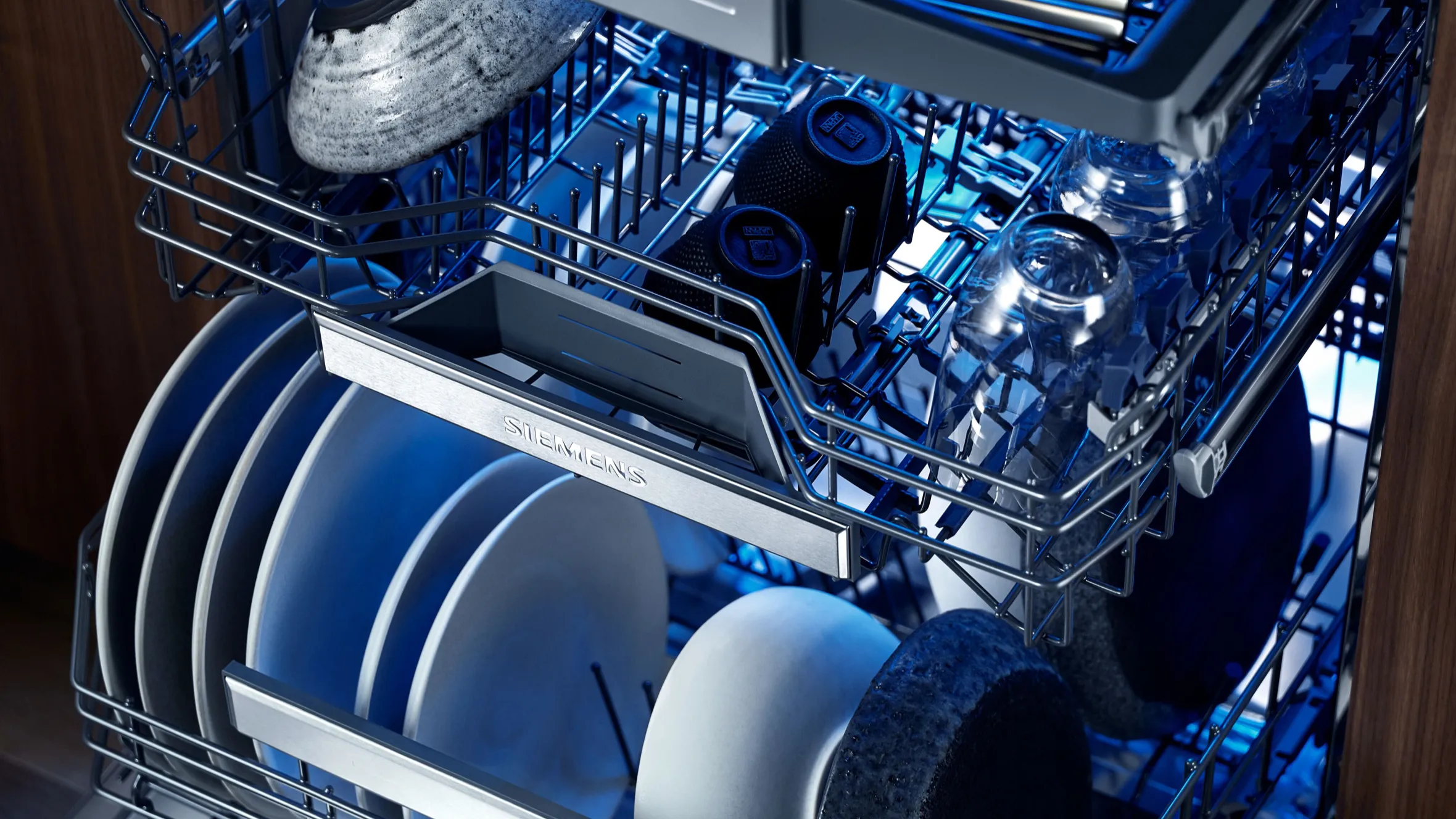Dishwasher interior with various dishes, glasses, and utensils arranged in a blue-tinted metal rack.