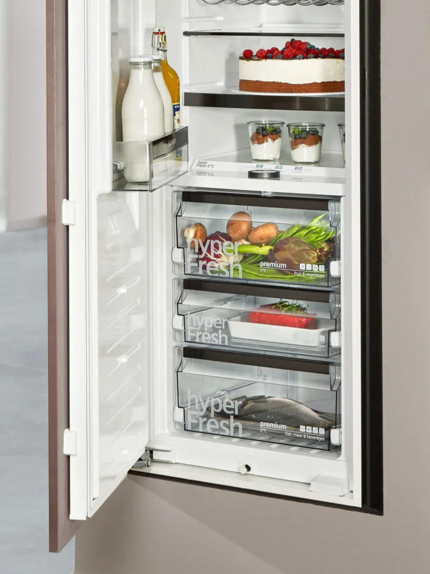 A close-up of the interior of a Siemens refrigerator, focusing on three transparent "hyperFresh" drawers filled with fresh food. The top drawer contains vegetables, the middle contains packaged meat, and the bottom holds a whole fish. Above the drawers, shelves are stocked with a cheesecake and desserts in glasses.