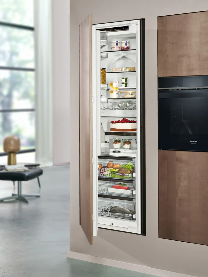 A tall, integrated Siemens refrigerator with a wood-paneled door, shown open in a modern living space. The well-stocked interior features multiple "hyperFresh" drawers and shelves holding fresh food and a cake.