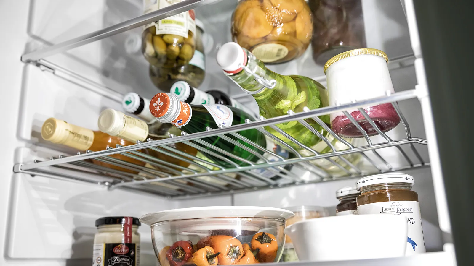 Open fridge filled with bottled drinks, jars, green apples, and containers with fruits and preserves on glass shelves.