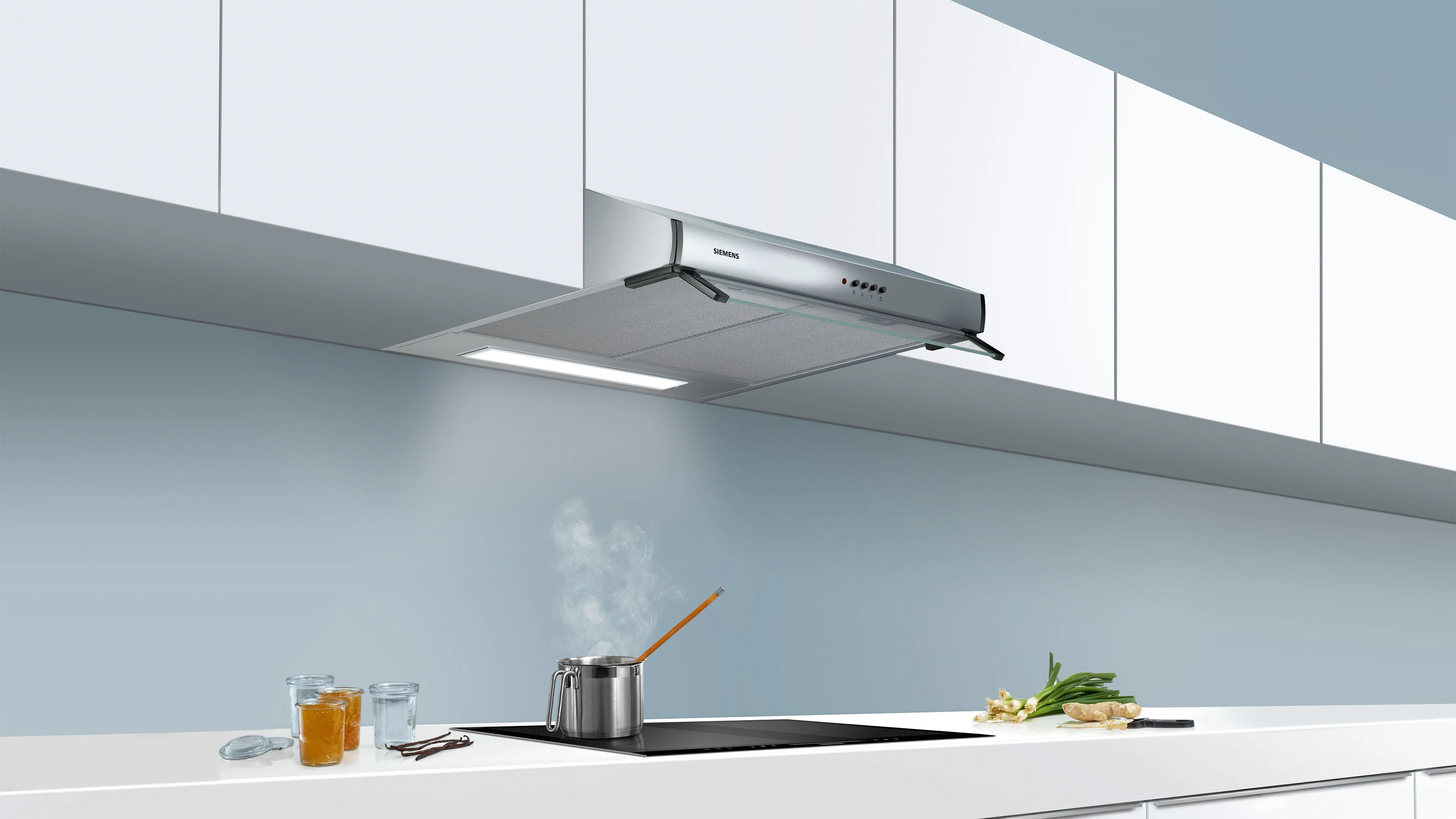 Modern kitchen with a silver hood above a stovetop, steaming pot, and glassware on a white countertop against a soft blue wall.