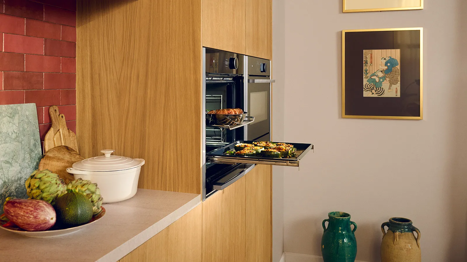 A modern kitchen features light wooden cabinetry and a red tiled backsplash. A sleek, black built-in oven has a baking tray filled with stuffed vegetables pulled out on its telescopic rails. Above it, another dish bakes in a higher rack. On the light-colored countertop to the left, a white Dutch oven sits next to a bowl of fresh produce including artichokes, eggplant, and avocado, with wooden cutting boards leaning against the wall. On the right, a light grey wall displays framed artwork and two decorative ceramic vases on the floor.