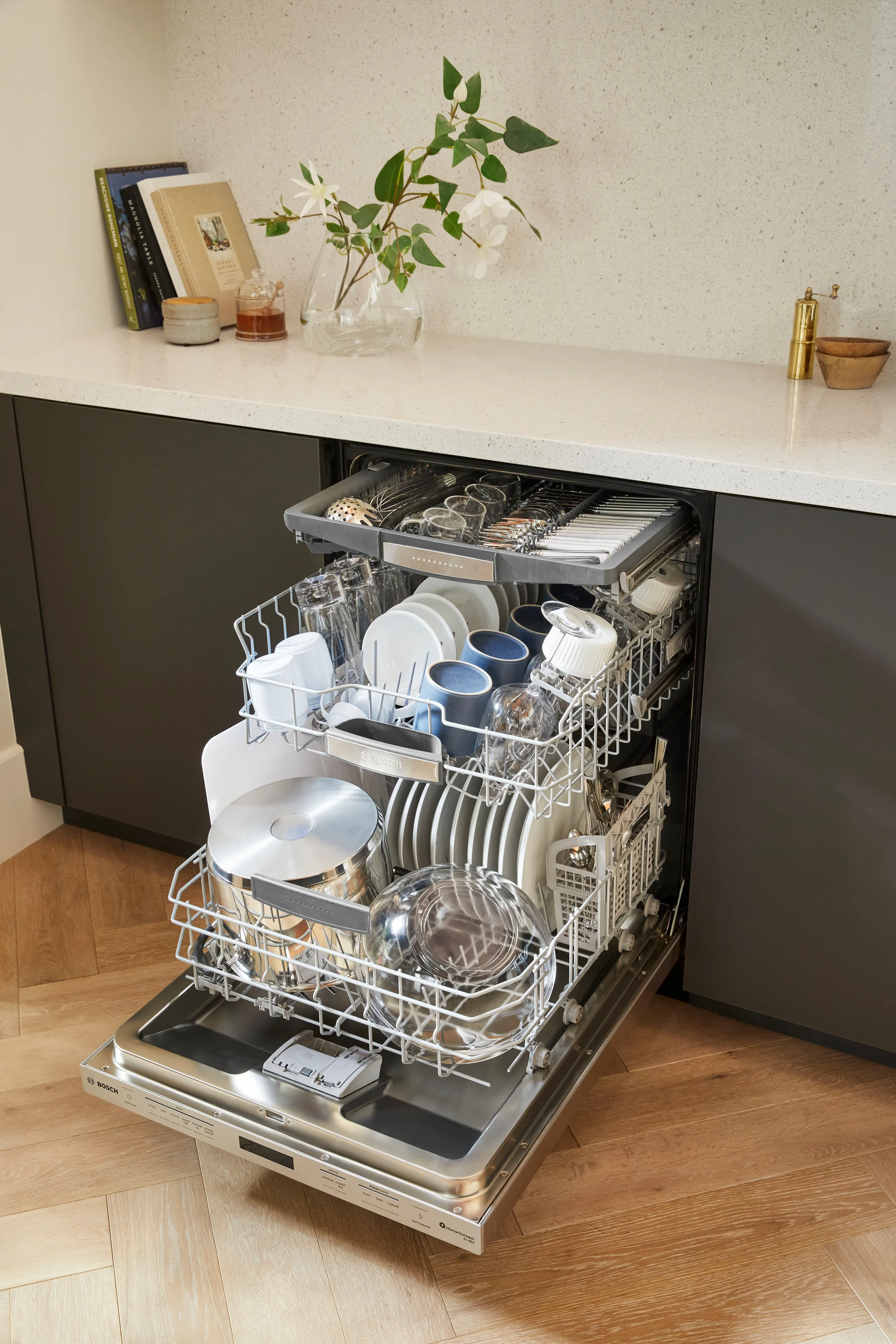 Open Bosch dishwasher with three racks full of clean dishes, utensils, and cookware in a modern kitchen setting.