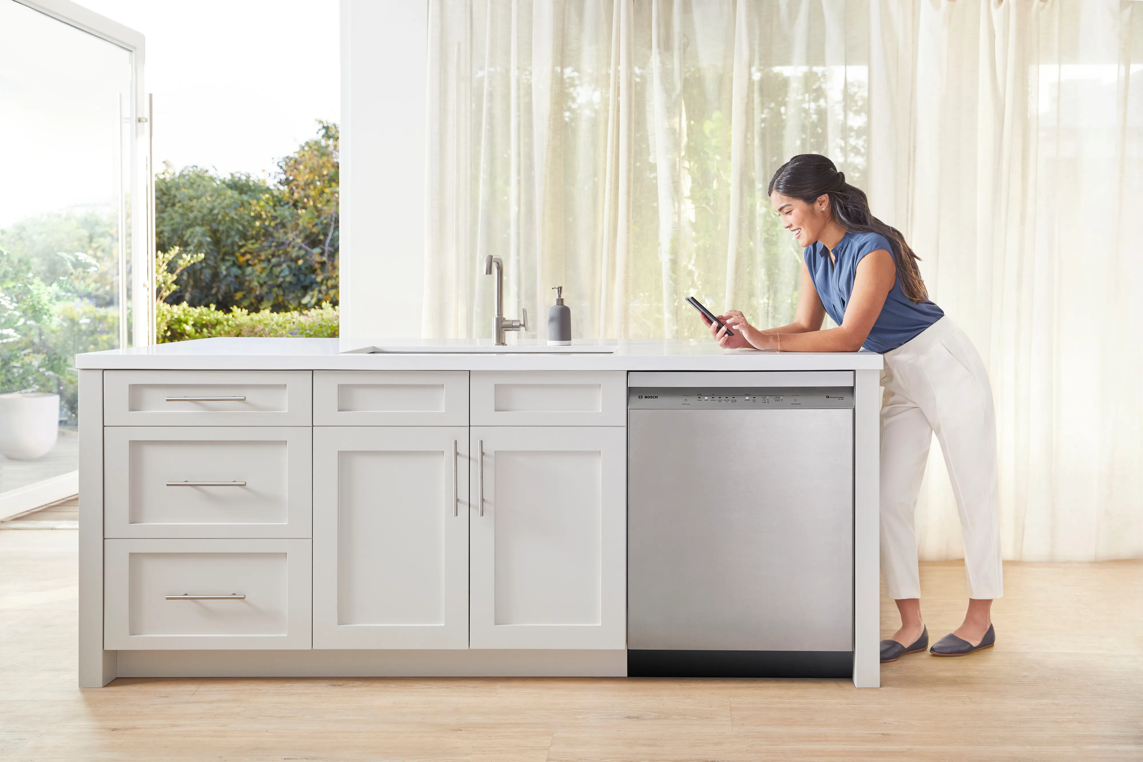 Woman using smartphone in modern kitchen with Bosch dishwasher and curtains.