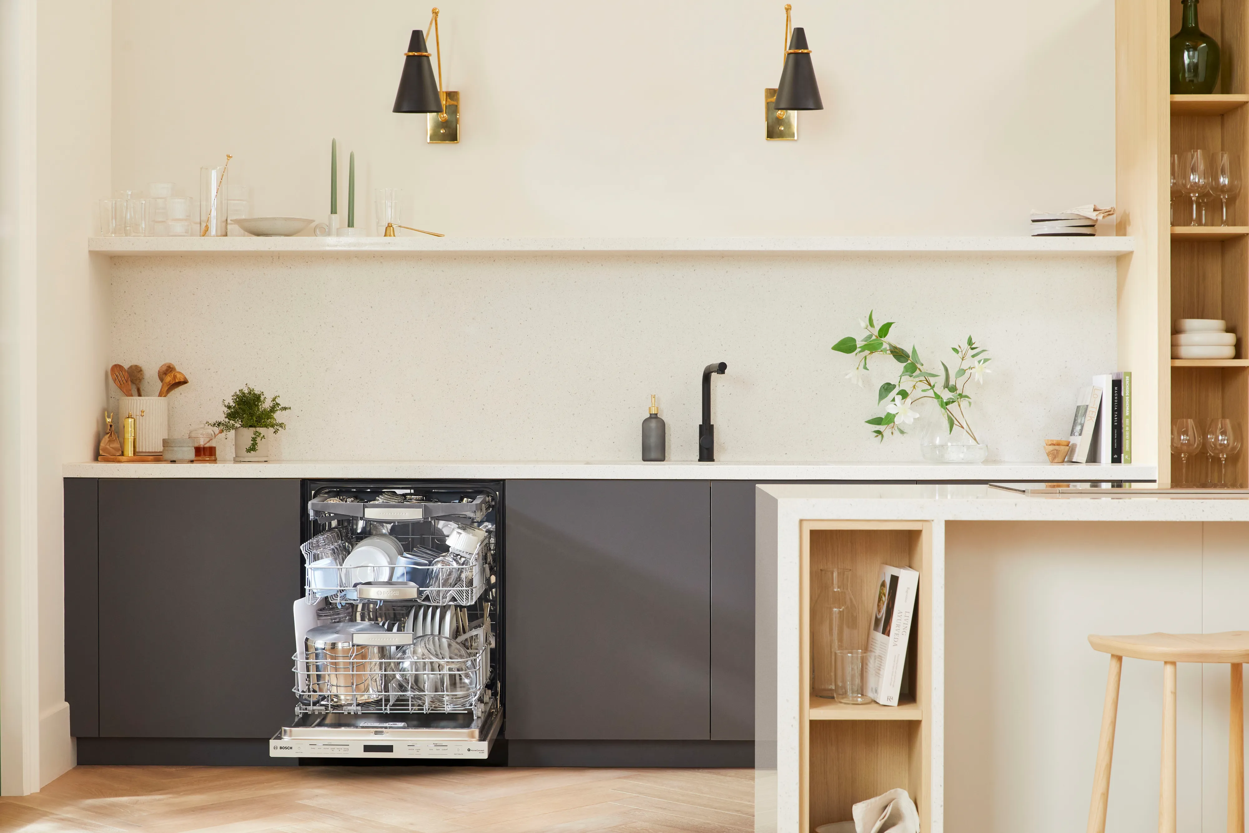 Modern kitchen with an open Bosch three rack dishwasher full of dishes under a white countertop and washed black shelves.