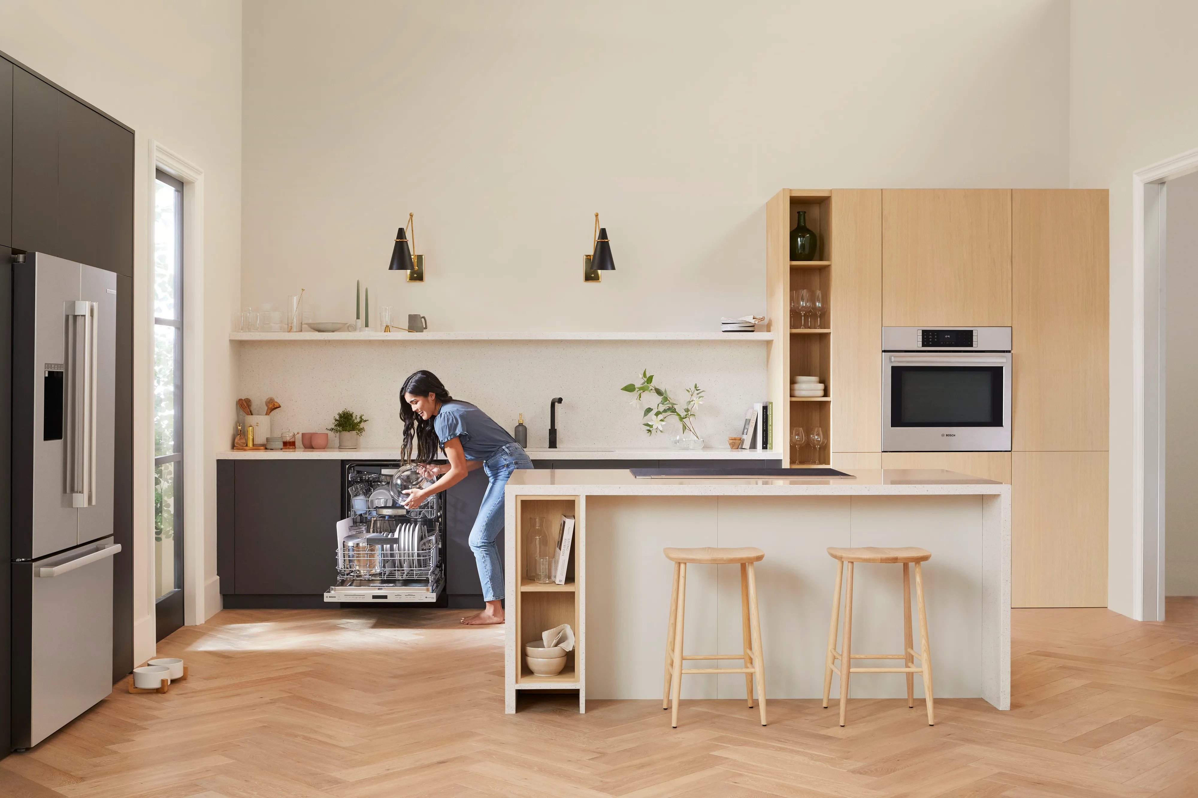 Modern kitchen with a woman loading a Bosch third rack dishwasher near island with stools and built-in oven.