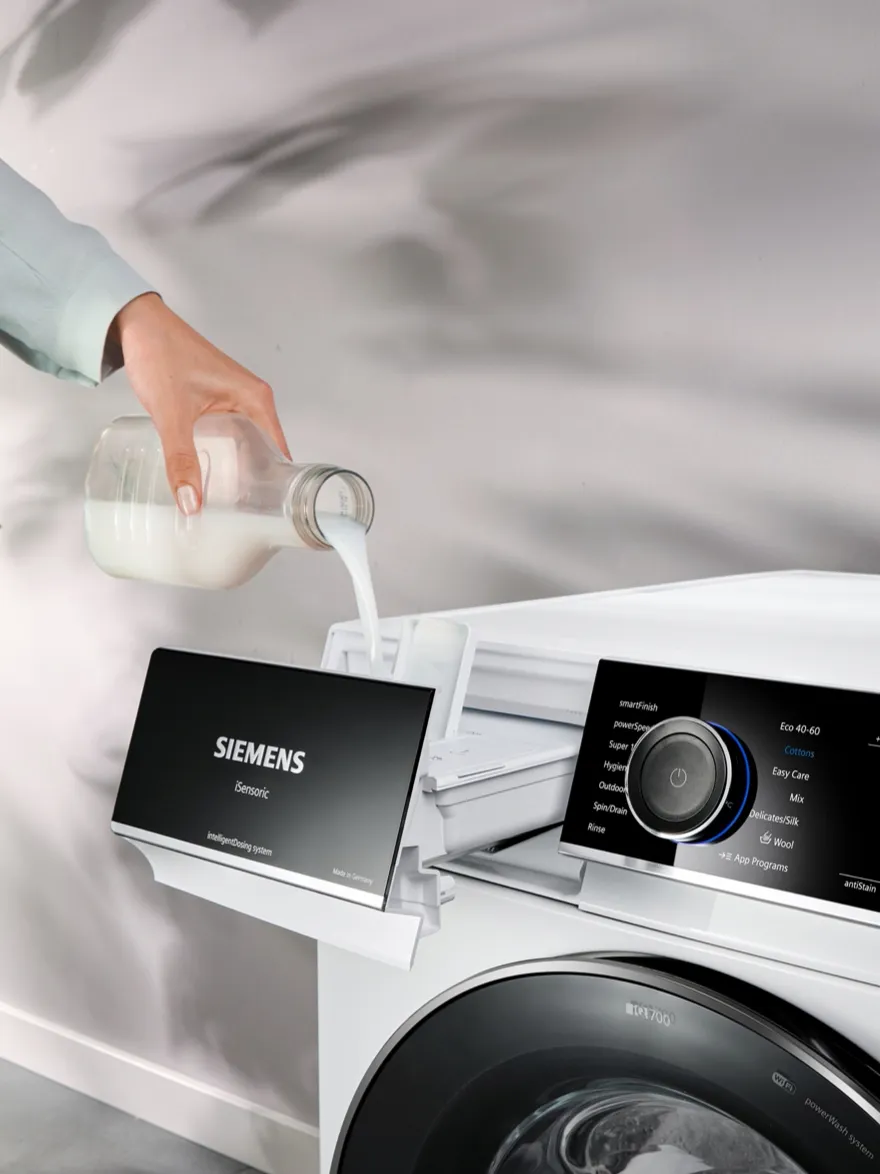 Washing machine with Siemens logo, hand pouring detergent, blurred background.
